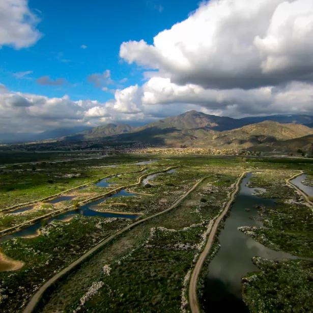 Aerial view of green valley with ponds/marshes and two-lane roads running through against backdrop of mountains and blue, cloud-covered sky