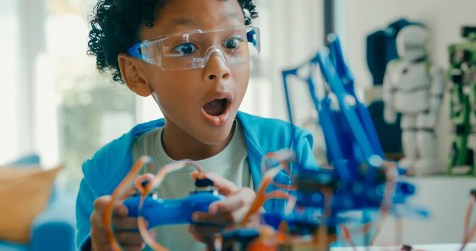 Closeup of Young African-American boy using remote control for robotics project amidst various technological tools and models at home.