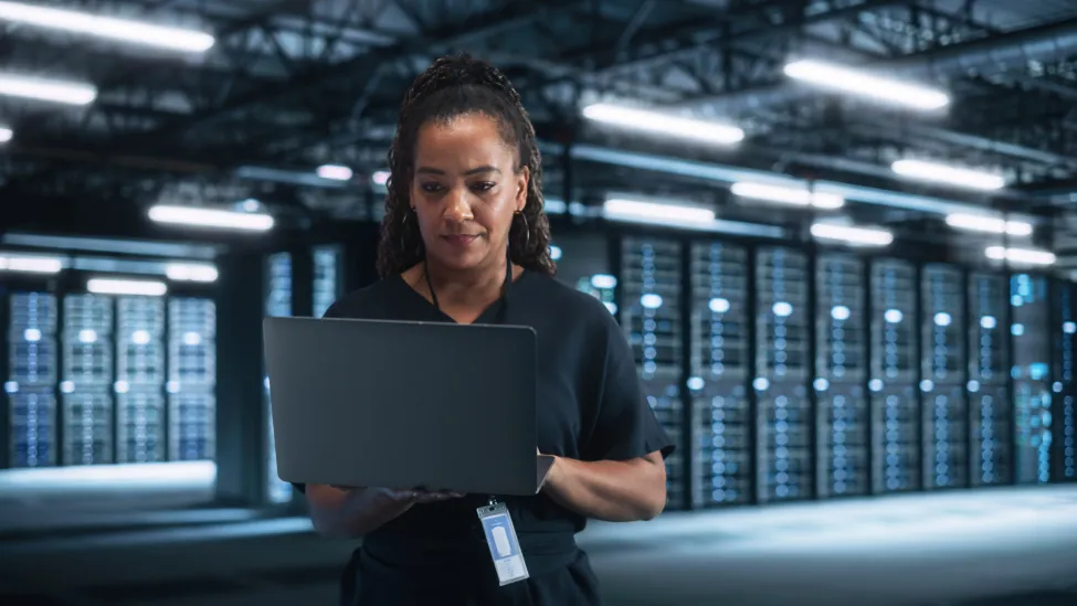 Successful Female IT Specialist Using Laptop Computer, while Standing at Big Warehouse Data Center. System Administrator working with Computing SAAS, Cloud Services. E-Business Digital Entrepreneur