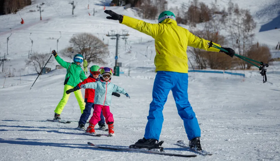 An instructor on a snowy mountain demonstrates skiing techniques to three children, all dressed in eye-catching winter gear on alpine resort
