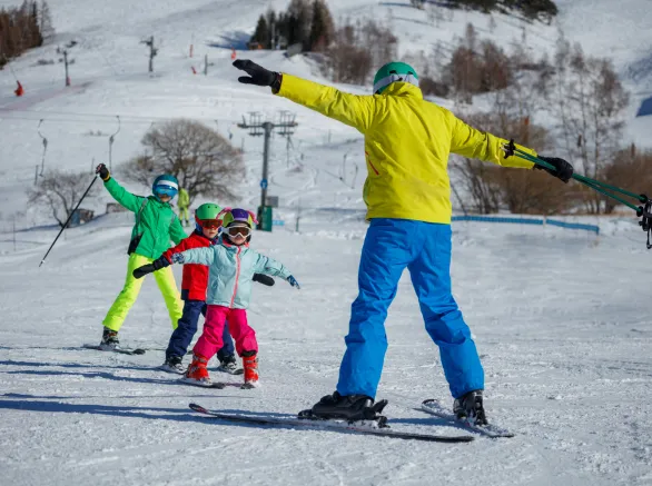 An instructor on a snowy mountain demonstrates skiing techniques to three children, all dressed in eye-catching winter gear on alpine resort