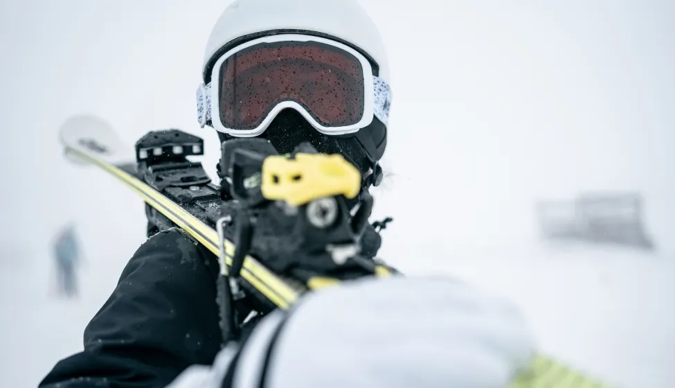 A close-up action shot of a skier wearing a white helmet and red-tinted goggles, gripping a yellow ski pole with a snowy, blurred background that conveys cold, movement, and outdoor sport.