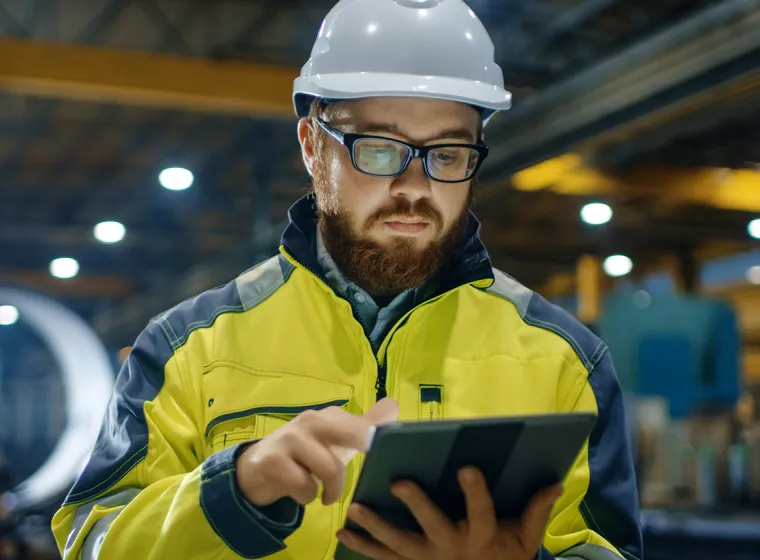 Inspector holds a clipboard and wears a hard hat in a factory setting. Exponent provides scientific and engineering risk assessments.
