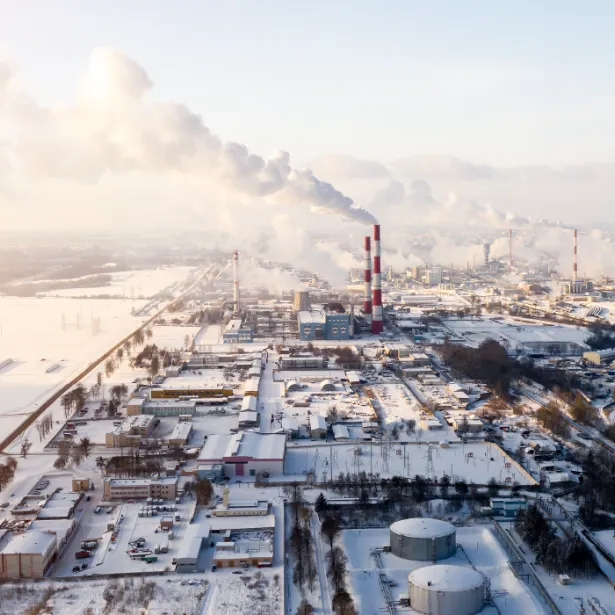 Aerial view of snow-covered industrial section of city with red and white striped smokestacks blowing smoke across hazy, dim-lit sky