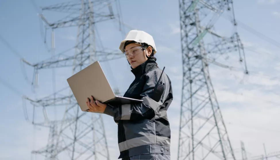 A utilities professional wearing goggles and helmet, staring at a laptop with 2 electrical towers in background